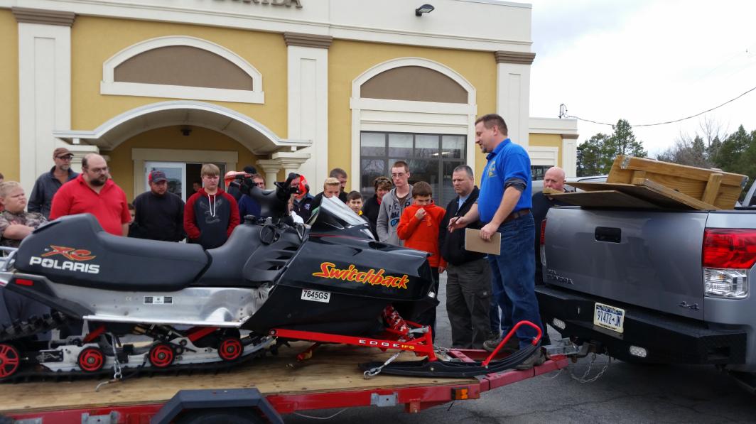 Instructor Lenz speaking to a group, pictured with a snowmobile on a trailer