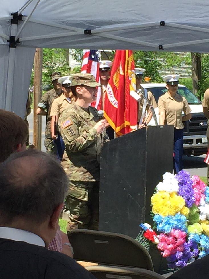 Guest speaker Sarah Secor-Jones speaks at a podium, with flowers in the foreground and flags in the background